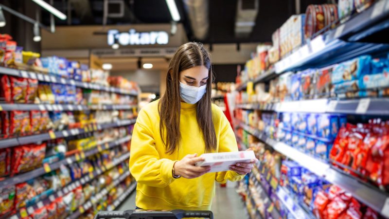 Social distancing in a supermarket. A young woman in a disposable face mask buying food and putting them in a grocery basket. Shopping during the Coronavirus Covid-19 epidemic